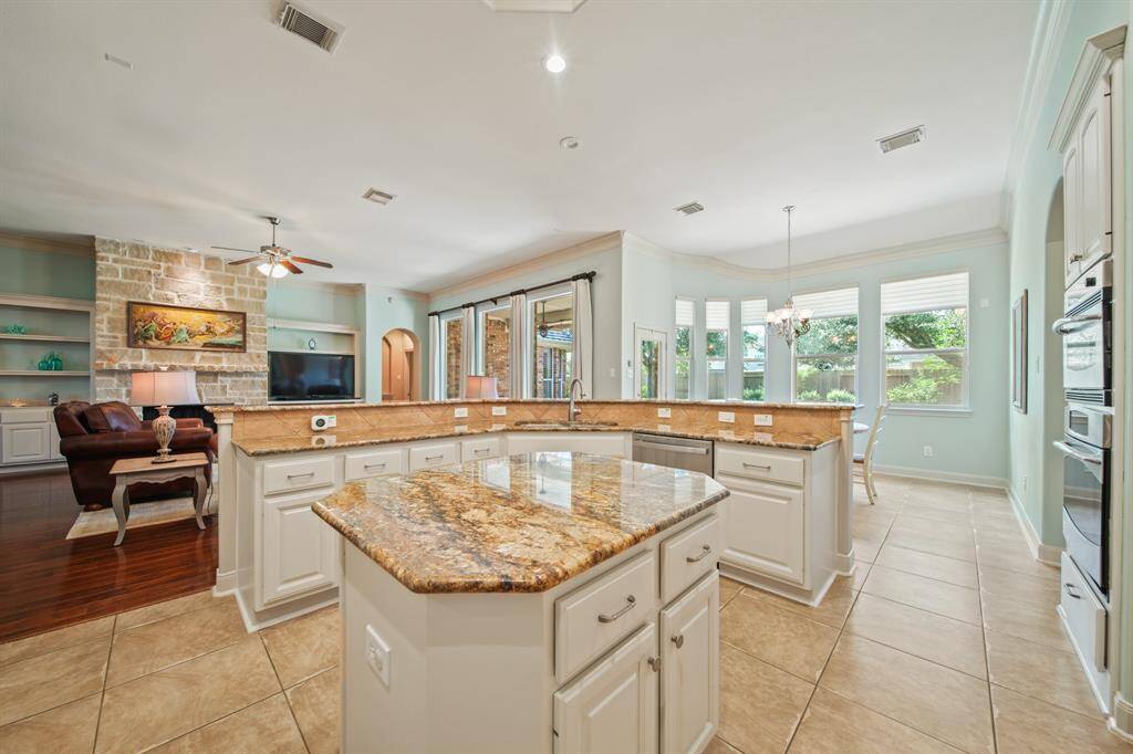 This spacious kitchen features a large granite island, ample cabinetry, and an open floor plan connecting to a cozy living area with a stone fireplace. Bright windows and a dining nook create a welcoming, airy atmosphere.