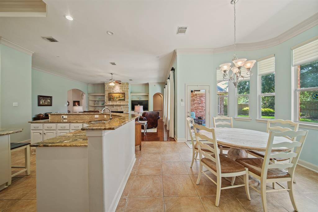 This spacious kitchen and dining area features a large island with granite countertops, ample natural light from multiple windows, and an open layout leading into a cozy living space with a stone fireplace. Perfect for entertaining and family gatherings.