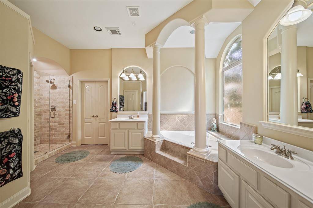 This spacious bathroom features a luxurious corner bathtub framed by elegant columns, a large walk-in shower with brick-style tiles, dual vanities, and ample natural light from a tall arched window, creating a serene and sophisticated atmosphere.