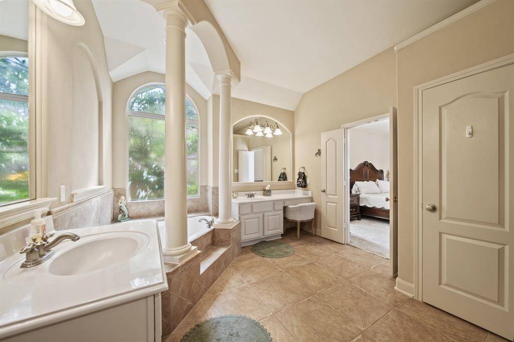 This spacious bathroom features elegant columns, a large soaking tub, and dual sinks. Natural light streams through arched windows, complementing the neutral tones. It leads directly into a cozy bedroom, offering convenience and luxury.