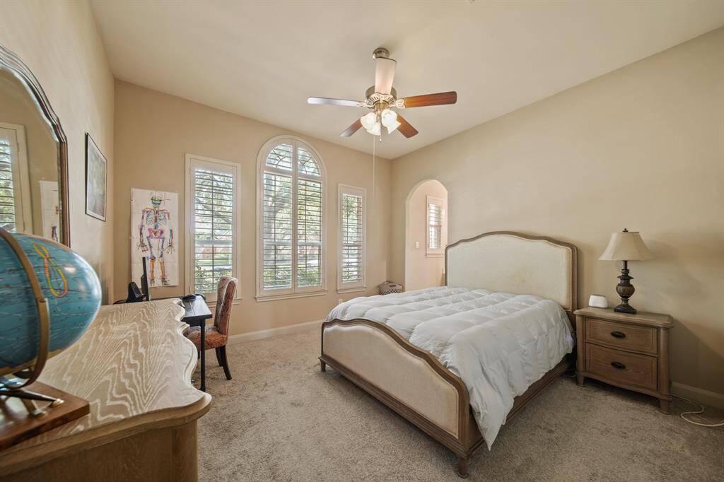 This cozy bedroom features soft beige walls and a ceiling fan. Natural light streams through large, arched windows with shutters. The room creates a functional and inviting space.