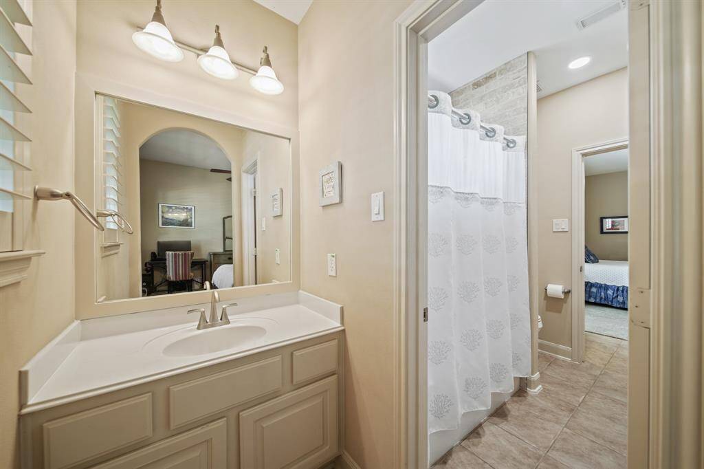 This photo features a bright bathroom with a large mirror, a vanity with ample storage, and a shower with a curtain. The neutral tones create a warm, inviting feel, and there's an adjacent bedroom visible through the doorway.