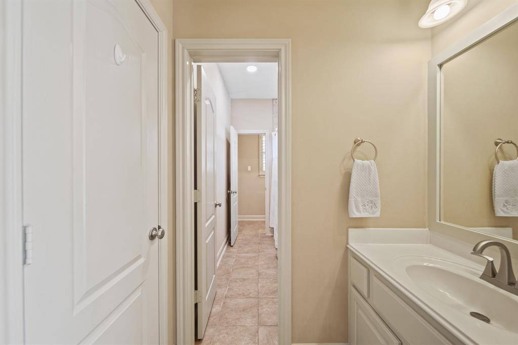 This photo shows the jack and jill bathroom with a single vanity, a large mirror, and a towel ring. It has tiled flooring and a neutral color palette, leading to the tub area and the adjoining bedroom.