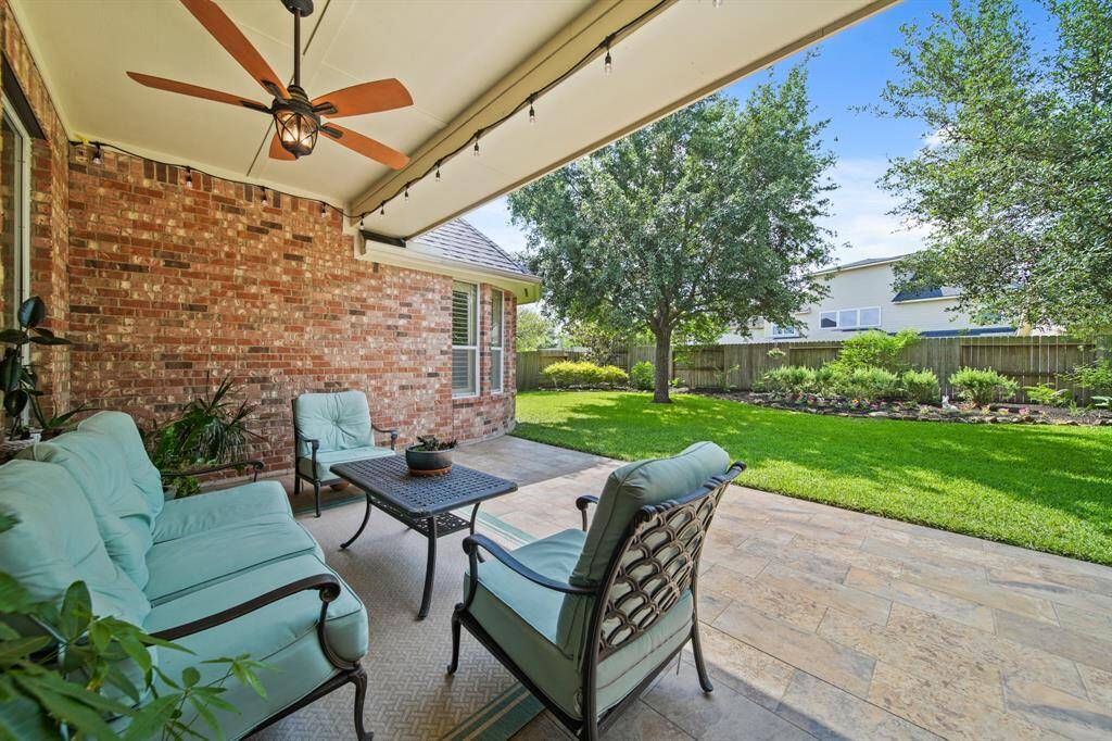This inviting patio features comfortable seating under a ceiling fan, set against a charming brick wall. It opens to a well-maintained backyard with lush grass, mature trees, and a landscaped border, providing a perfect space for relaxation and outdoor entertaining.