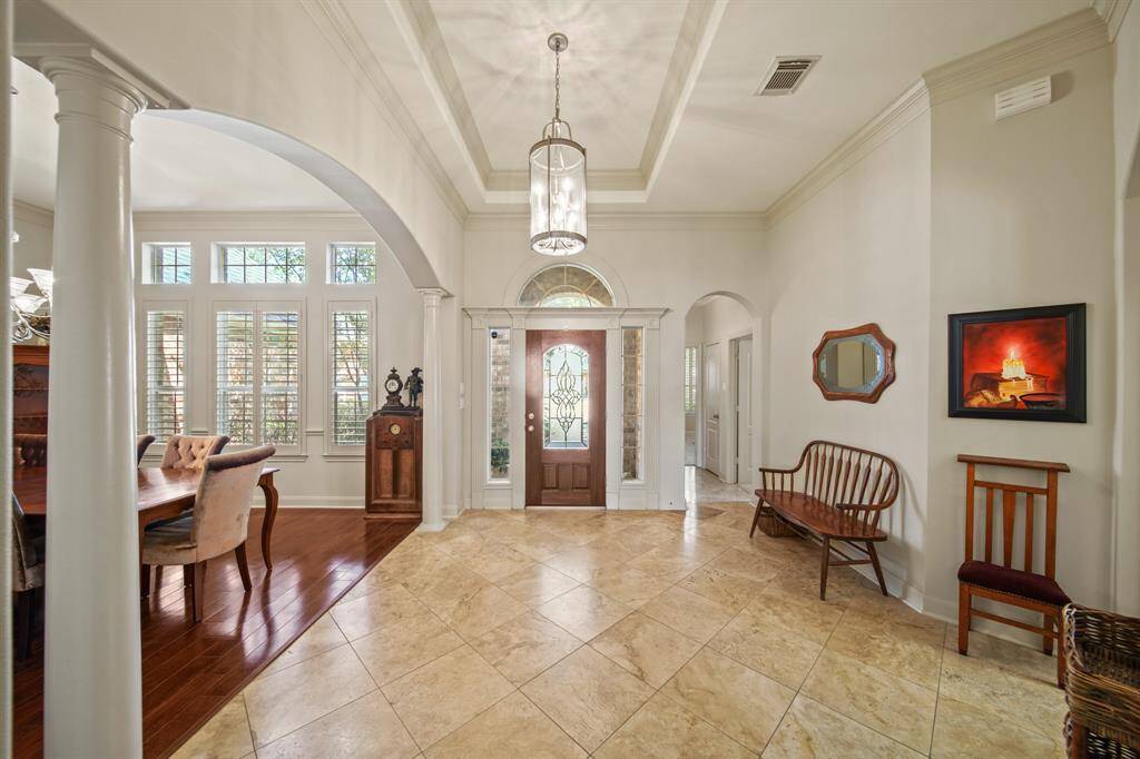 This elegant entryway features high ceilings, a decorative chandelier, and large windows with abundant natural light. The space is complemented by polished tile flooring and classic columns, leading into a dining area with rich wood accents.