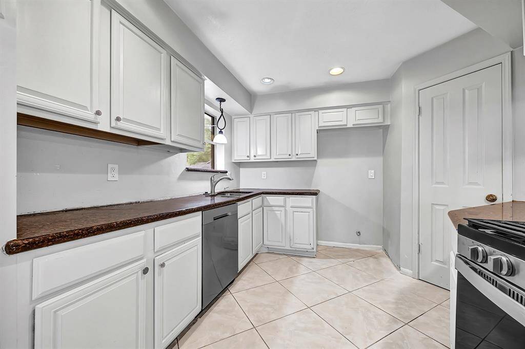 Bright, spacious kitchen with white cabinetry, dark countertops, and stainless steel appliances. Features a large window with natural light, recessed lighting, and neutral tile flooring.