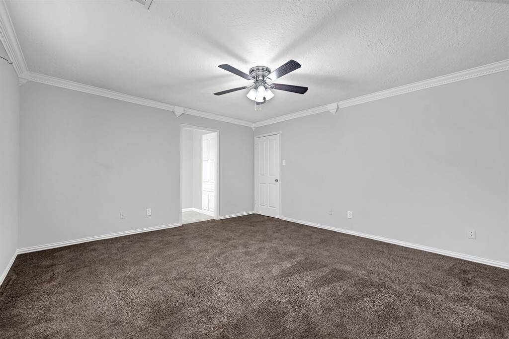 This spacious room features neutral walls, a dark carpet, and a ceiling fan with lights. It includes a white door leading to another space, offering versatility and comfort.