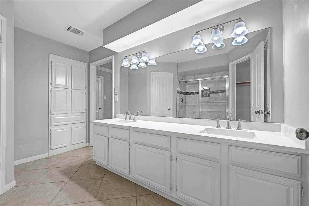 Spacious bathroom featuring a double vanity with sleek fixtures, a large mirror, modern lighting, and neutral tile flooring. Ample storage and a walk-in shower add to the functionality.