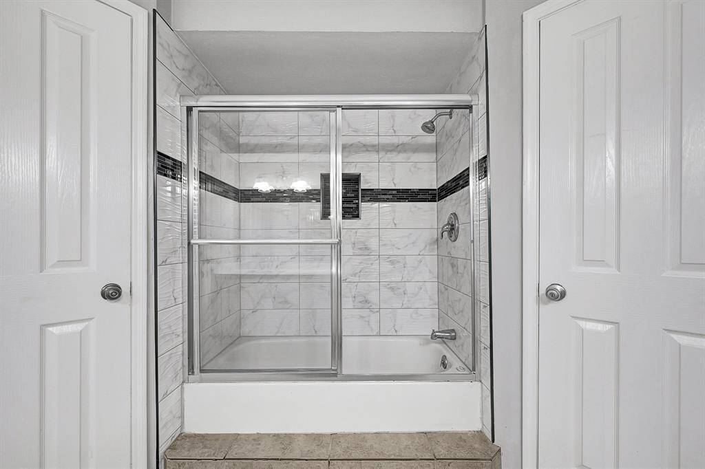 This photo features a modern bathroom with a glass-enclosed shower and bathtub combination. The walls are tiled with a marble-like finish, accented by a horizontal dark tile strip. The space is bright and clean, with two white doors on either side.