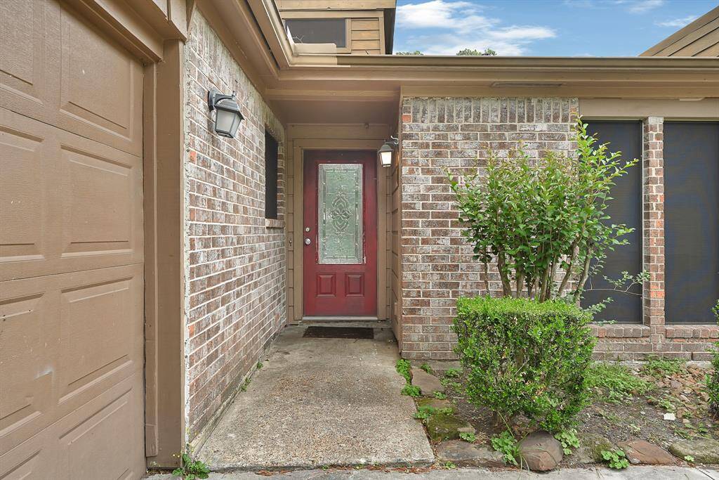 This photo shows the entrance of a home with a red door and decorative glass panel. It's flanked by brick walls and a garage on the left. The path is bordered by a neatly trimmed shrub, and the exterior has a classic, inviting look.