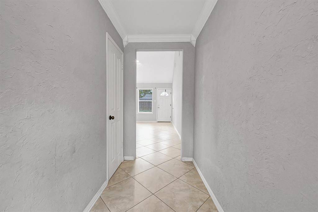 Bright hallway with textured gray walls, crown molding, and light tile flooring leading to a well-lit room with large windows and a front door.