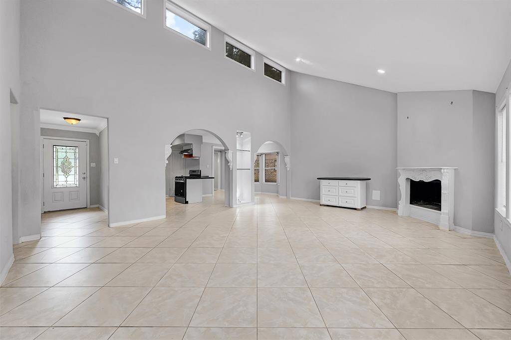 This spacious living area features high ceilings and ample natural light from upper windows. It includes elegant tile flooring, a decorative fireplace, and arched entryways leading to the kitchen. The open layout offers versatility for various furniture arrangements.