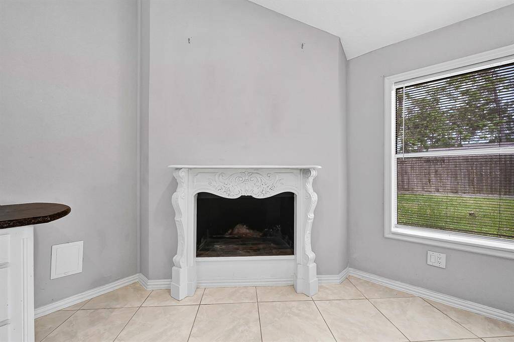 Cozy corner of a room featuring a decorative white fireplace with intricate detailing. The space is brightened by a large window with blinds, offering a view of the backyard. The neutral tile flooring and light gray walls create a welcoming ambiance.