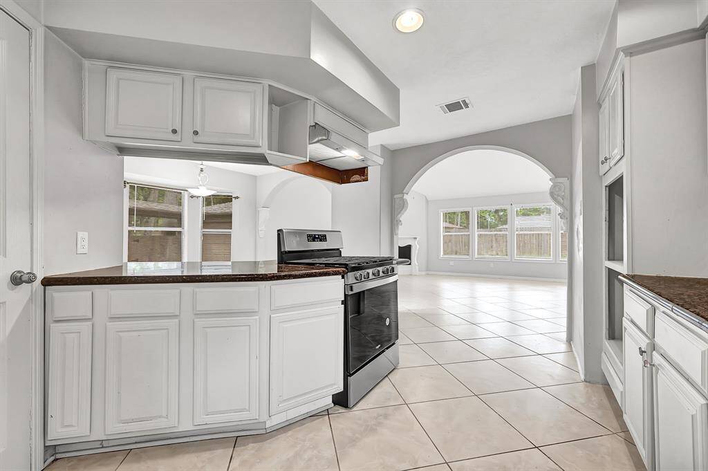 This photo showcases a spacious, bright kitchen with white cabinetry, a modern black stove, and tiled flooring. It opens up to a large living area with an arched entryway and ample natural light from multiple large windows.