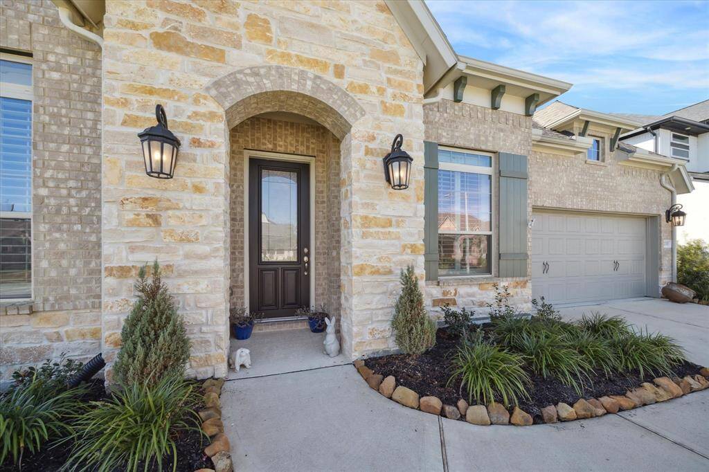This photo showcases the charming front entrance of a house, featuring a stone facade with an arched entryway, and attractive landscaping. Flanking lantern-style lights add a welcoming touch, and a spacious driveway leads to a 2 ½ -car garage.
