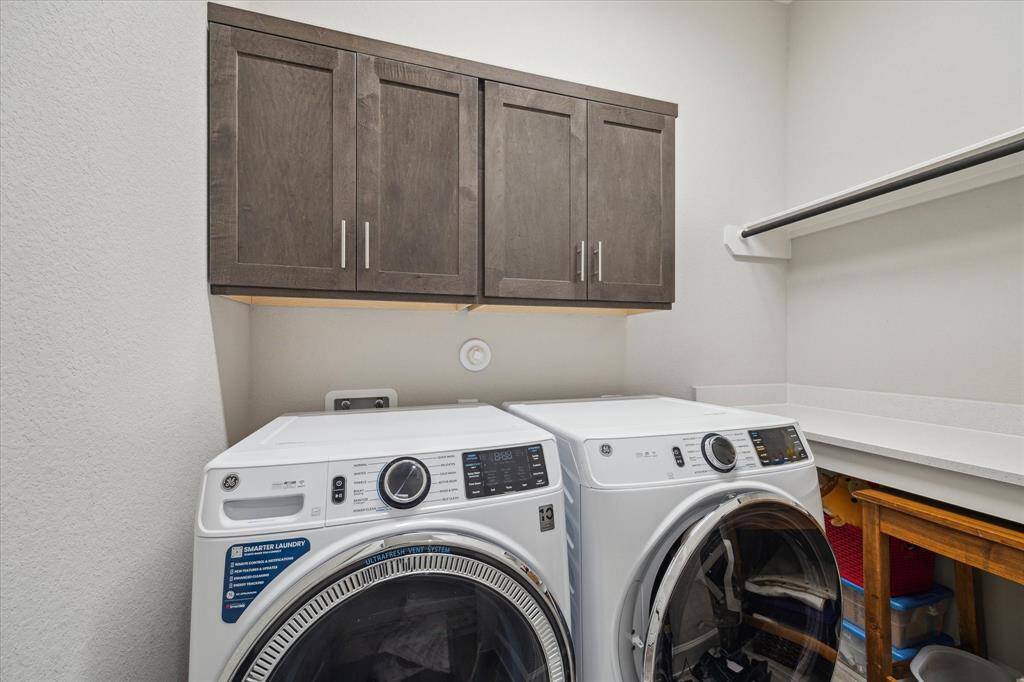 The well-designed laundry room features upper cabinets for additional storage, a countertop for folding, and space for a full-size washer and dryer. It also includes a hanging rod and additional shelving for enhanced convenience and organization.