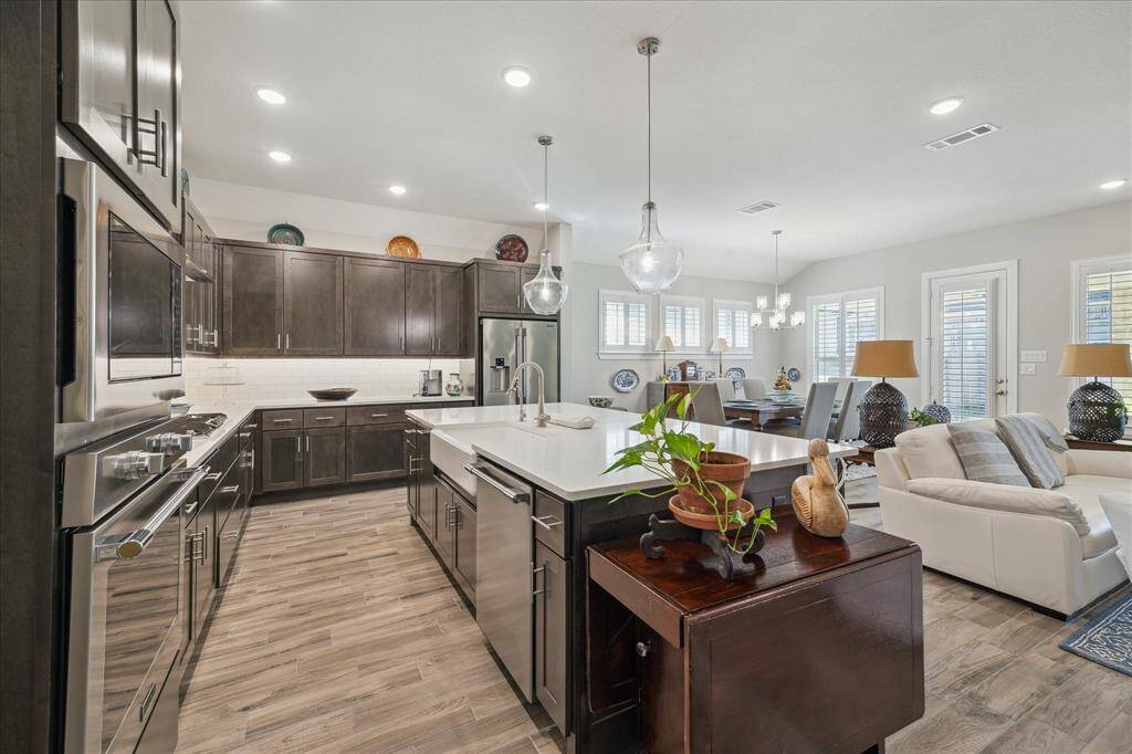 A chef's dream, the gourmet kitchen features a spacious island with seating and stainless steel appliances showcasing an open-concept kitchen and living area. The adjacent living space includes comfortable seating with enough room for a piano, large windows, and a dining area with a contemporary chandelier, creating a bright and inviting atmosphere.