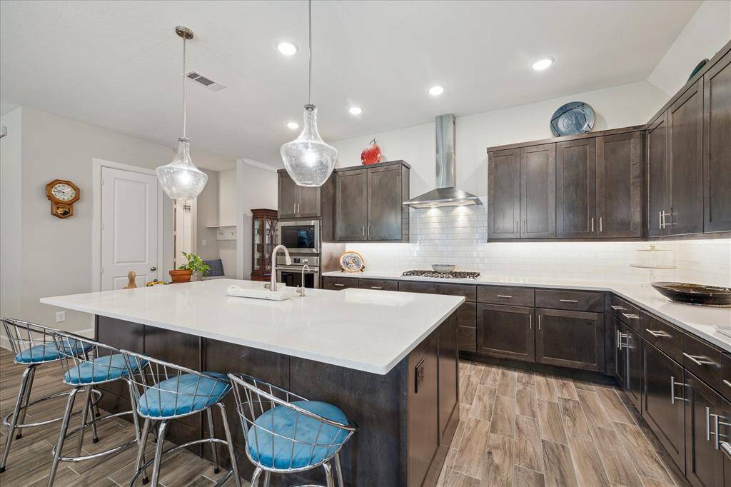 This modern kitchen features sleek dark cabinetry, stainless steel appliances, and a large central island with seating. It offers ample lighting with pendant fixtures and recessed lights, and the open layout seamlessly connects to the adjacent hallway and living areas.