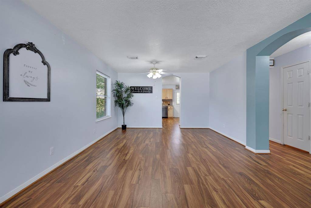 This photo showcases a spacious, open-concept living area with hardwood floors, neutral walls, and ample natural light. The room features a ceiling fan, decorative wall accents, and leads into a modern kitchen through an arched doorway.