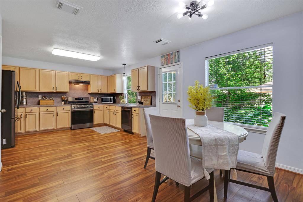 This bright kitchen features wood flooring, light wood cabinetry, and modern appliances. A cozy dining area is by a large window, offering natural light and a view of greenery.