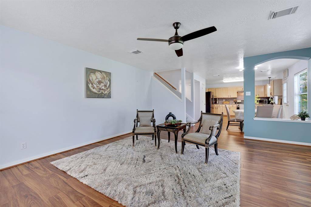 This inviting family room features vinyl plank floors and an open layout leading to a modern kitchen. Natural light flows through large windows, enhancing the airy feel.