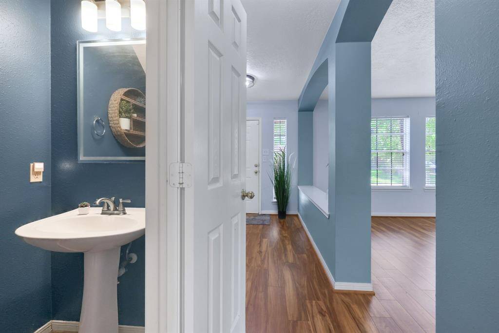 This photo features a bright entryway with wood flooring and a view into a cozy powder room with a pedestal sink. The space is enhanced by large windows for natural light and an elegant color scheme.