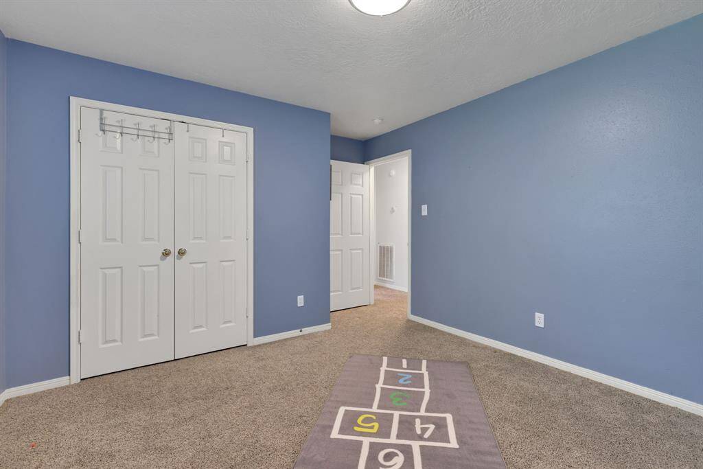 This room features soft blue walls, a cozy carpet, and a spacious closet with double doors. It has a simple overhead light fixture and a playful hopscotch rug, making it ideal for a child's bedroom or a versatile space.