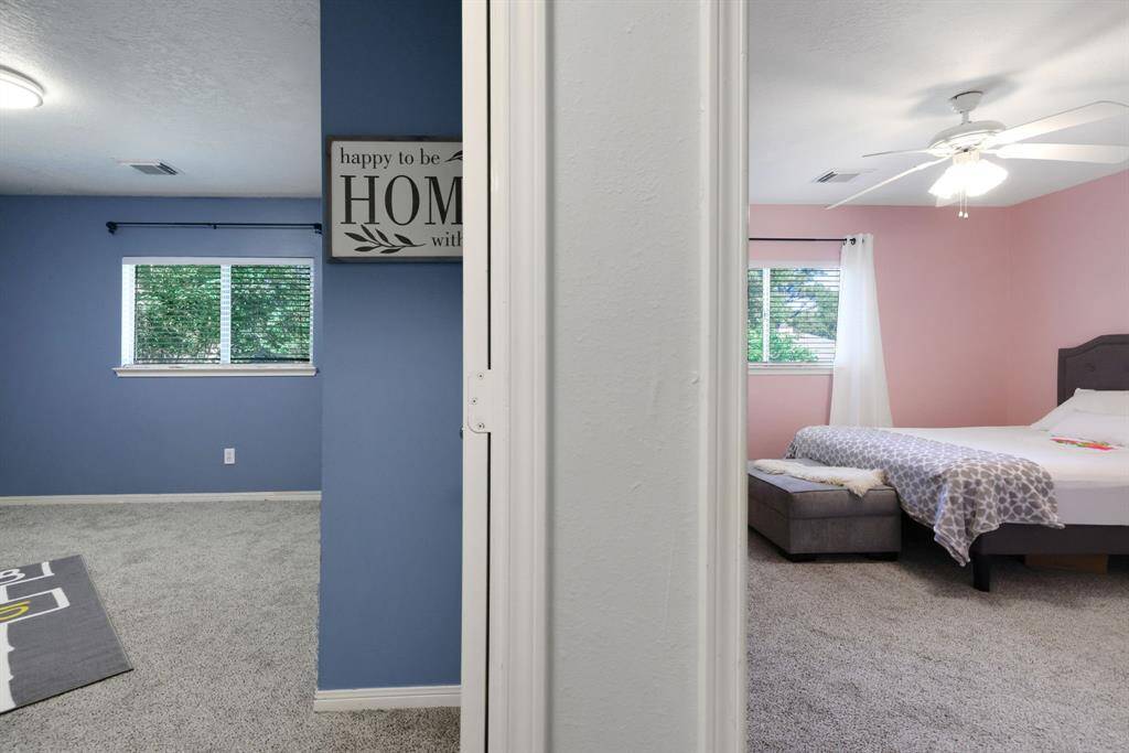 This photo shows two adjacent rooms divided by a central wall. The left room has blue walls and a window with blinds, while the right room features pink walls, a ceiling fan, and a cozy bed setup. Both rooms have carpeted floors, creating a comfortable and inviting atmosphere.