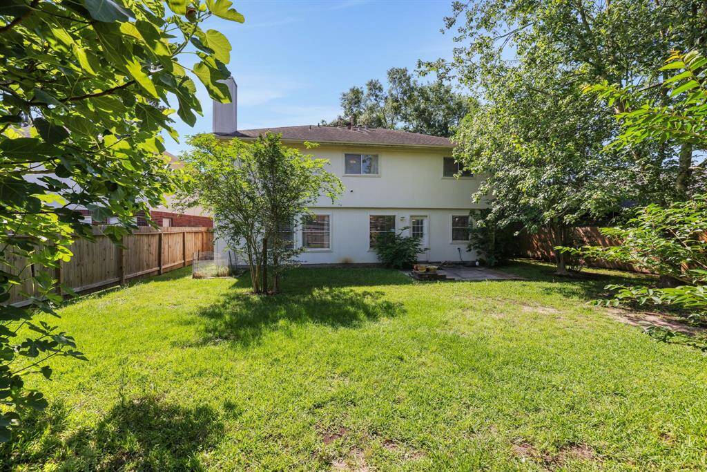 This photo showcases a spacious backyard with lush grass and mature trees providing shade. The two-story house features a light exterior and multiple windows, offering a view to the green space. A wooden fence encloses the area, creating a private and serene outdoor setting.