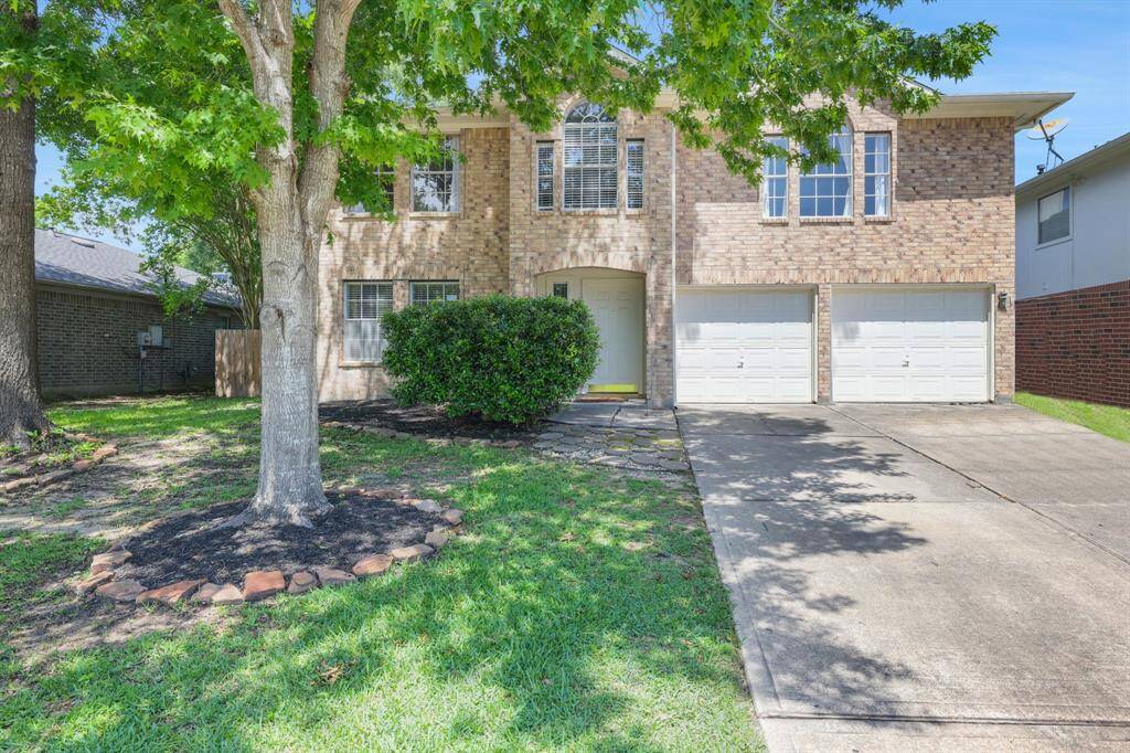 This is a two-story brick home with a double garage, set on a well-maintained lot with mature trees and a tidy lawn. The driveway provides ample parking, and the front entrance is welcoming with a landscaped pathway.
