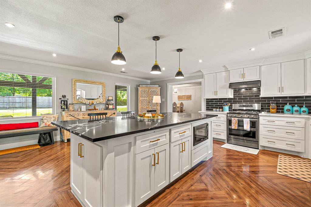 This kitchen features a spacious open layout with a large island and sleek black quartz. It includes modern appliances, white cabinetry, and stylish pendant lighting. The wood flooring adds warmth, and large windows provide ample natural light.