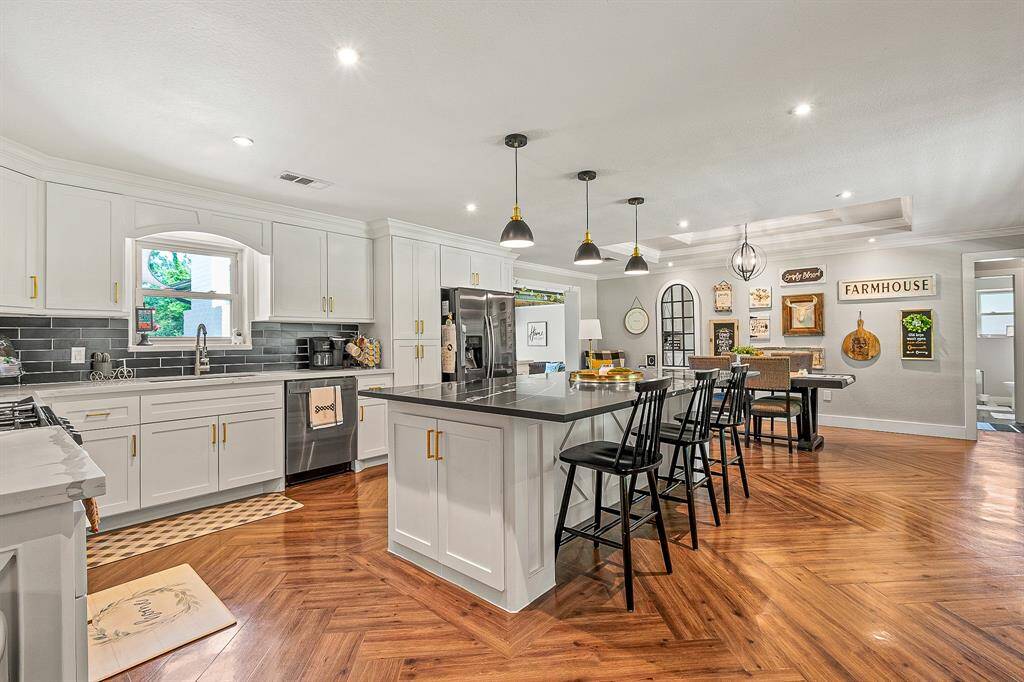 This spacious kitchen features modern cabinetry with gold accents, a large central island with seating, stainless steel appliances, and hardwood floors. The open design connects to a dining area with stylish decor, creating a warm and inviting atmosphere.