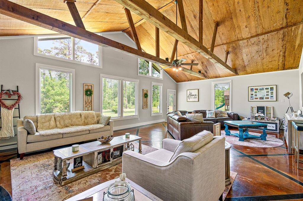 This spacious living room features a vaulted wood ceiling with exposed beams, large windows offering plenty of natural light, and a mix of cozy seating. The open layout has a warm, inviting atmosphere, perfect for relaxation and entertaining.