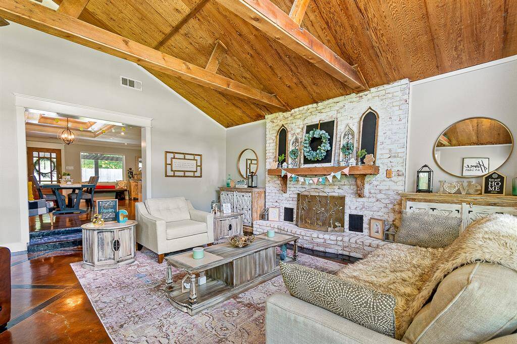 This cozy living room features a high wooden ceiling with exposed beams, a rustic brick fireplace, and tasteful decor. A large window offers plenty of natural light, while the open layout connects to a charming dining area. Perfect for relaxation and entertaining.