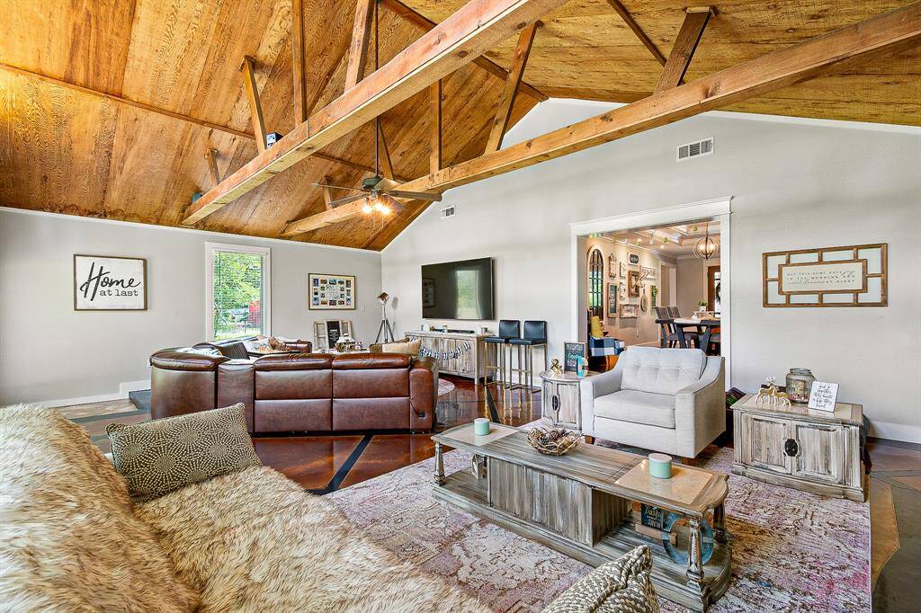 This spacious living room features a vaulted wooden ceiling with exposed beams, creating a warm and inviting atmosphere. The space is well-lit with natural light from large windows, and there's an open view into the dining area, enhancing the home's open-concept feel.