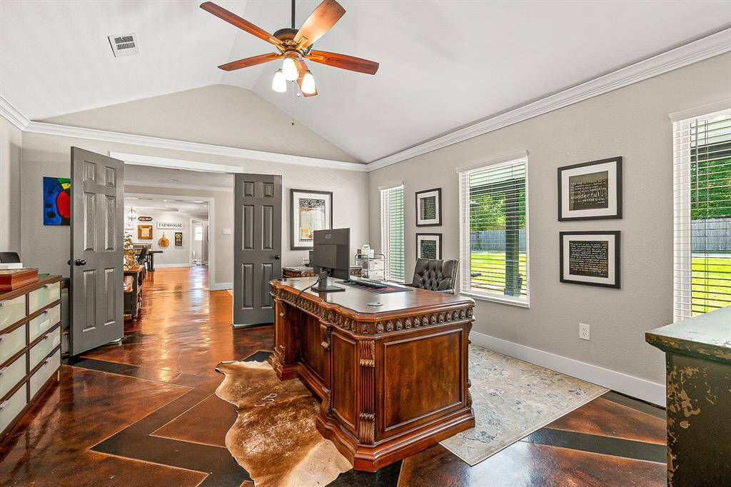 This photo showcases a spacious home office with a vaulted ceiling. It has ample natural light from multiple windows, modern ceiling fan, and attractive dark doors leading to an open area. Perfect for a productive workspace.