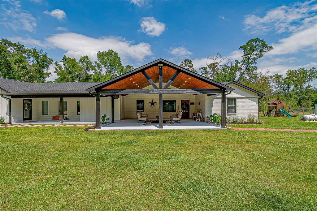 This photo features the large covered patio perfect for outdoor relaxation. The house has a modern, single-story layout with a light exterior and dark roof. The yard includes a play area, offering a great family-friendly space surrounded by greenery.