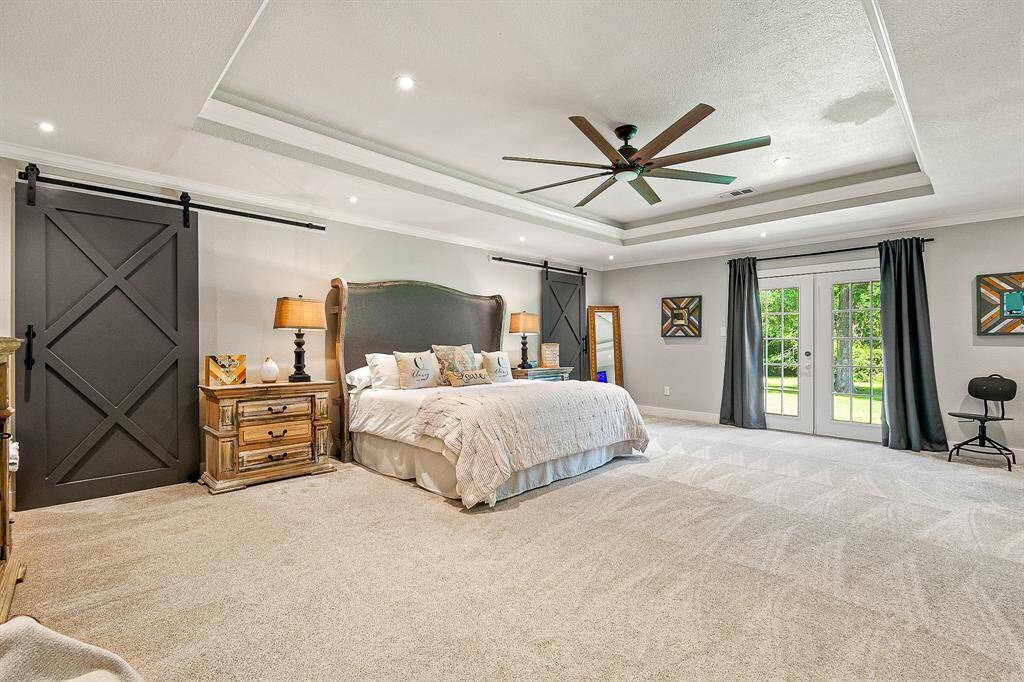 This spacious bedroom features a stylish tray ceiling with recessed lighting, and a central ceiling fan with ample natural light from French doors leading to outside.