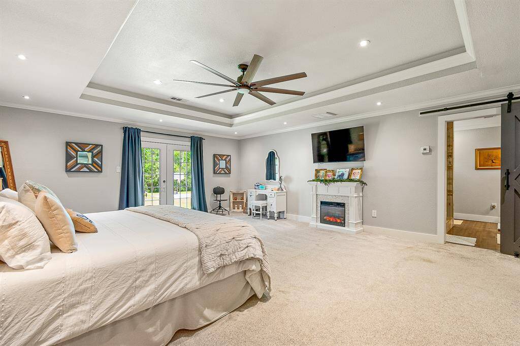 This spacious bedroom features a tray ceiling with recessed lighting and a ceiling fan, offering a modern and airy feel. The neutral carpet and walls create a calming atmosphere, while a sliding barn door adds a rustic touch to the connected closet.
