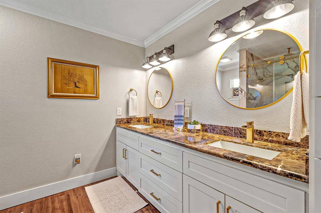 This primary bathroom features a double vanity with a luxurious marble countertop, gold fixtures, and two round mirrors. It has modern lighting, white cabinetry, and a warm wood floor, creating a stylish and functional space.