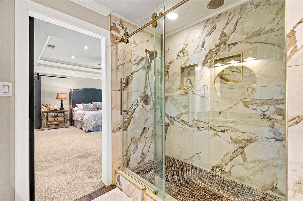This photo showcases a luxurious bathroom featuring a glass-enclosed shower with elegant tiles and gold accents. Adjacent is the cozy primary bedroom and a classic design, visible through an open doorway.