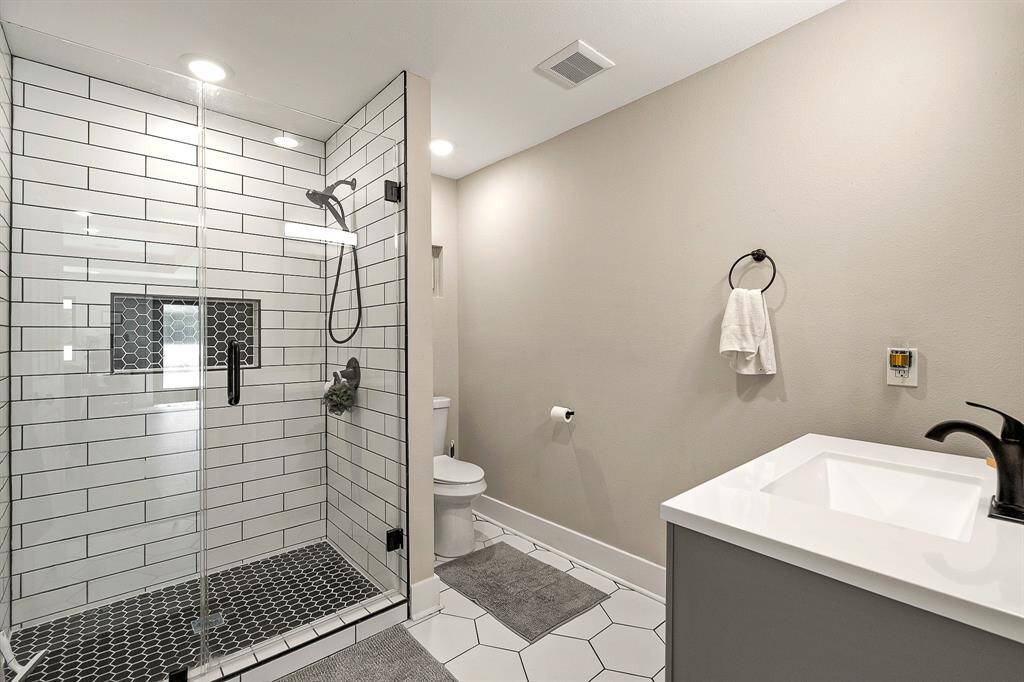 Guest house bathroom has a spacious glass-enclosed shower featuring white subway tiles and a sleek black hexagonal floor. It includes a contemporary vanity, large mirror, and neutral wall colors, creating a clean and stylish look.