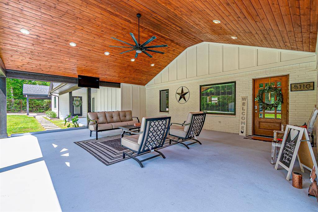 This photo showcases a spacious, covered outdoor patio with a wood-paneled ceiling and ceiling fan. It features comfortable seating, a wall-mounted TV, and decorative elements, creating an inviting space for relaxation and entertainment. The area overlooks a lush green yard, enhancing its appeal for outdoor living.