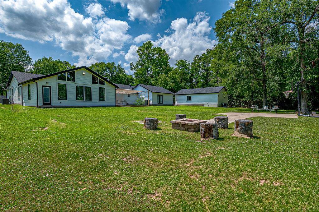 This photo showcases a modern single-story home with large windows and a spacious, well-maintained lawn. The backyard features a cozy fire pit area with tree stump seating, surrounded by lush greenery and mature trees, providing a serene outdoor space.