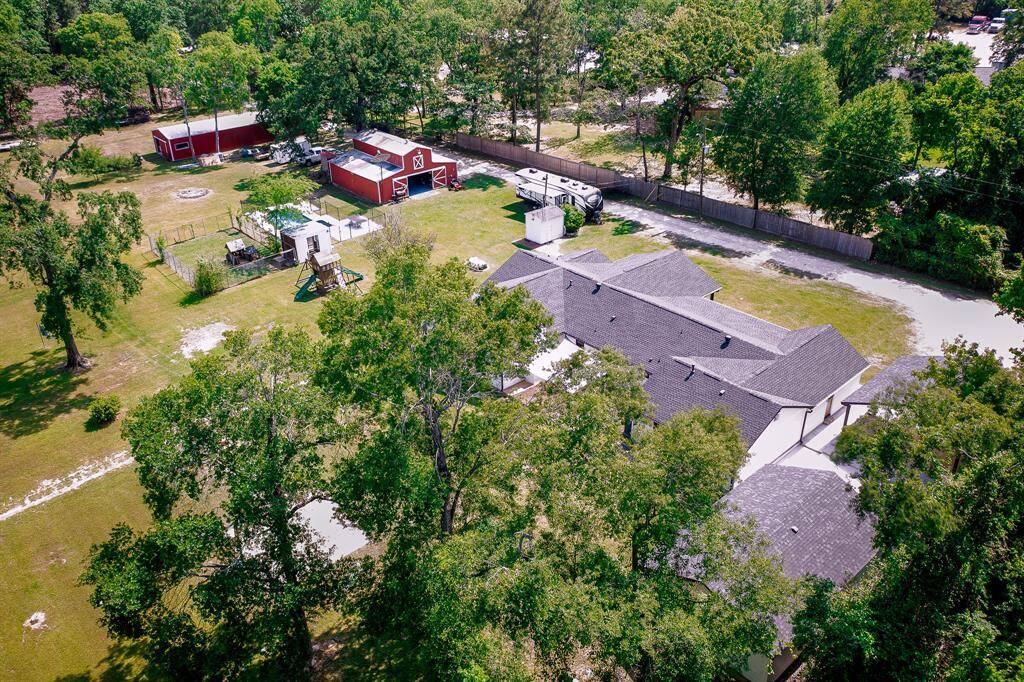 This aerial view showcases a spacious property with a large house surrounded by lush trees. The backyard features a play area, fenced sections and the side of the lot has 2 pole barns offering ample space for outdoor activities and storage.