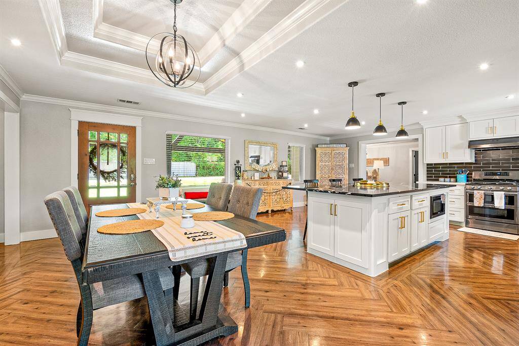 This photo showcases a spacious, open-concept kitchen and dining area with elegant wood flooring. The kitchen features a large island with modern pendant lighting, white cabinetry, and stainless steel appliances. The dining area is accented by a stylish chandelier and ample natural light from large windows, creating a warm and inviting atmosphere.