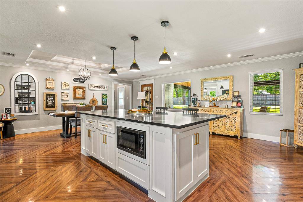 This spacious kitchen features a large island with a built-in microwave, modern pendant lighting, and elegant wood flooring. The open layout connects to a cozy dining area, with stylish decor and ample natural light from large windows showcasing the outdoor view.