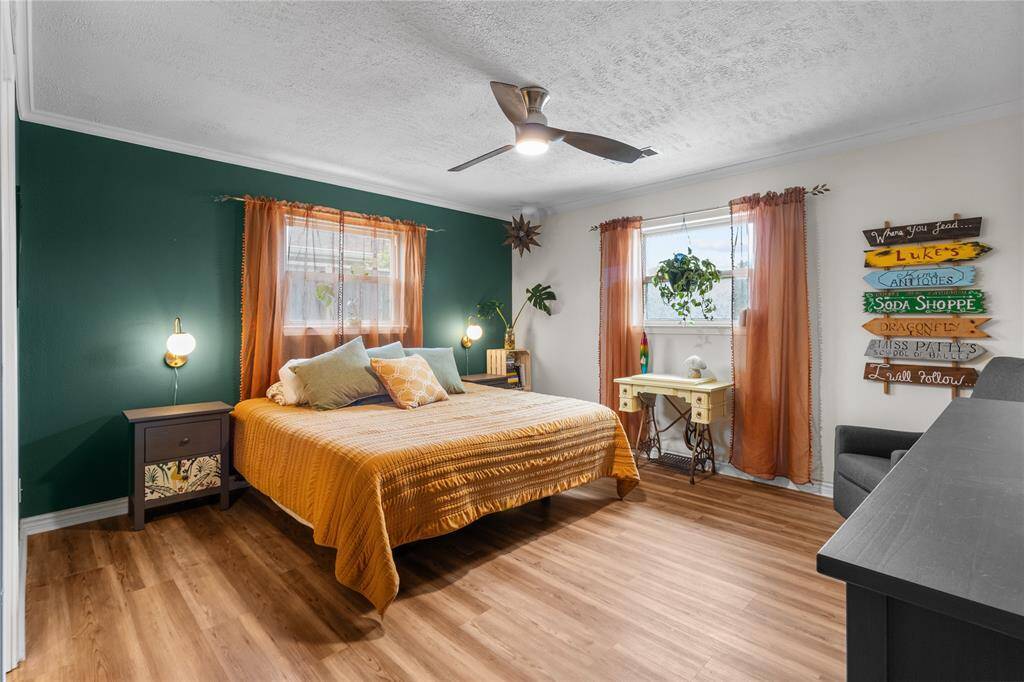 Bright and cozy downstairs primary bedroom with a modern ceiling fan, wood flooring, and a vibrant accent wall. Features include two large window.  Ideal for relaxation.