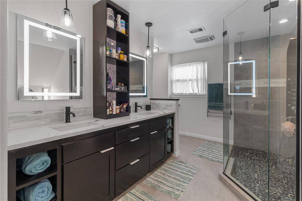 This modern bathroom features a sleek double vanity with ample storage, illuminated mirrors, and stylish pendant lighting. It boasts a spacious glass-enclosed shower with contemporary fixtures and a pebble stone floor. The space is brightened by a large window, creating a clean and inviting atmosphere.