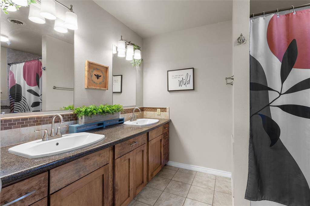 This bathroom features a modern design with dual sinks set in a granite countertop, wooden cabinetry, and contemporary lighting. The neutral tones are complemented by decorative backsplash and LED lighting.